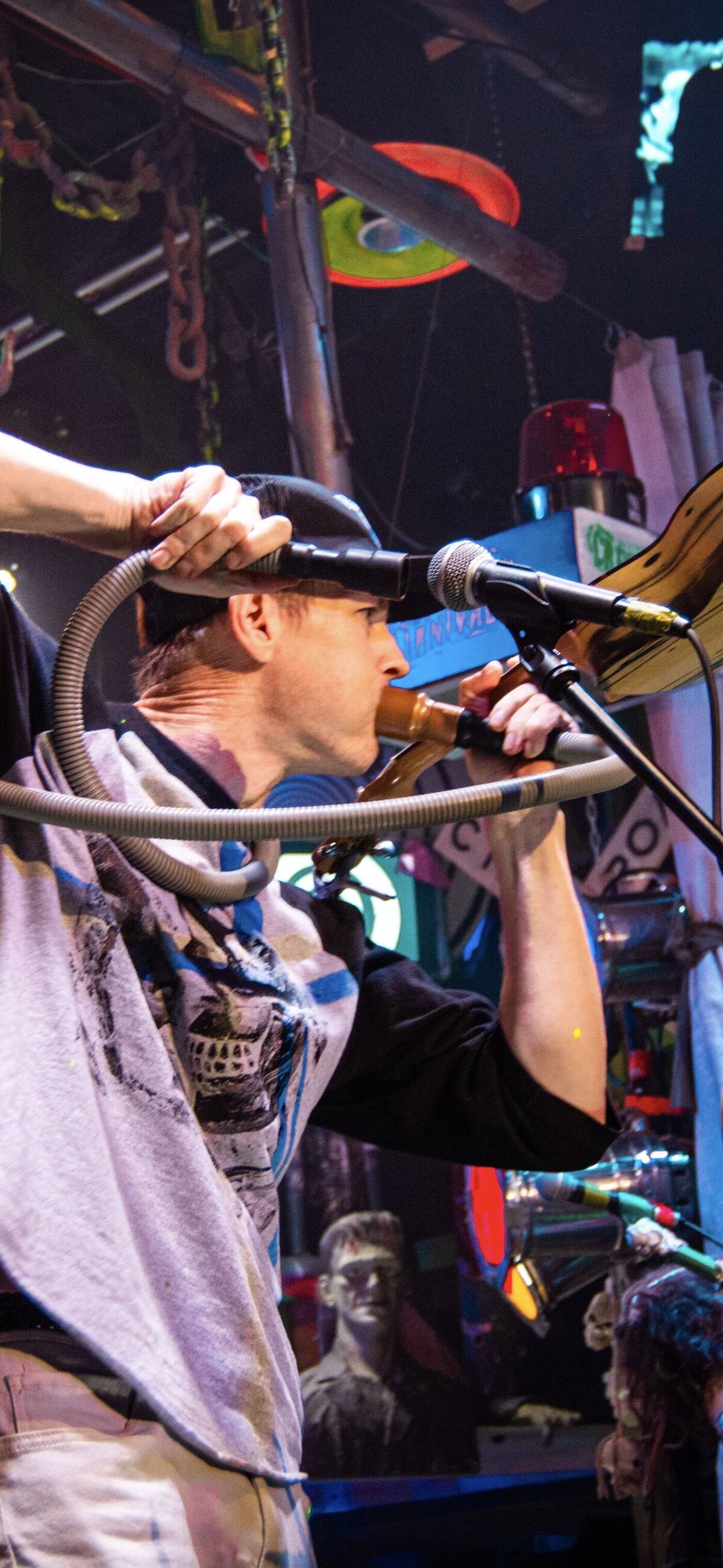 Ryan onstage at Creepsville Carnival playing a ukulele at a microphone, surrounded by chandeliers, hypnosis spirals, and haunt decor.