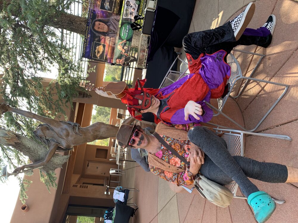 Ryan in a red jester mask and red-and-purple jester outfit reclining in a chair next to a friend in a mustache and sunglasses, in a Sedona plaza.