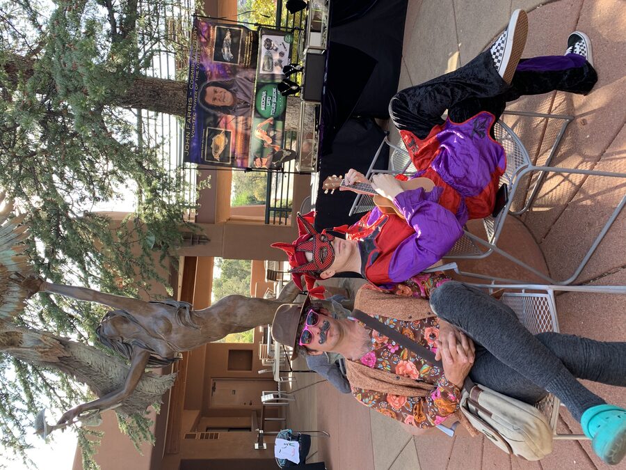 Ryan in the same jester mask and costume sitting with the same friend, now holding up a ukulele in mid-gesture.