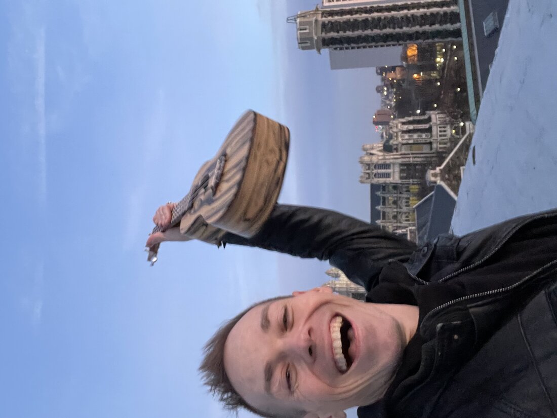 Ryan on a rooftop at dusk holding a ukulele up in the air, laughing, with the city skyline behind him.
