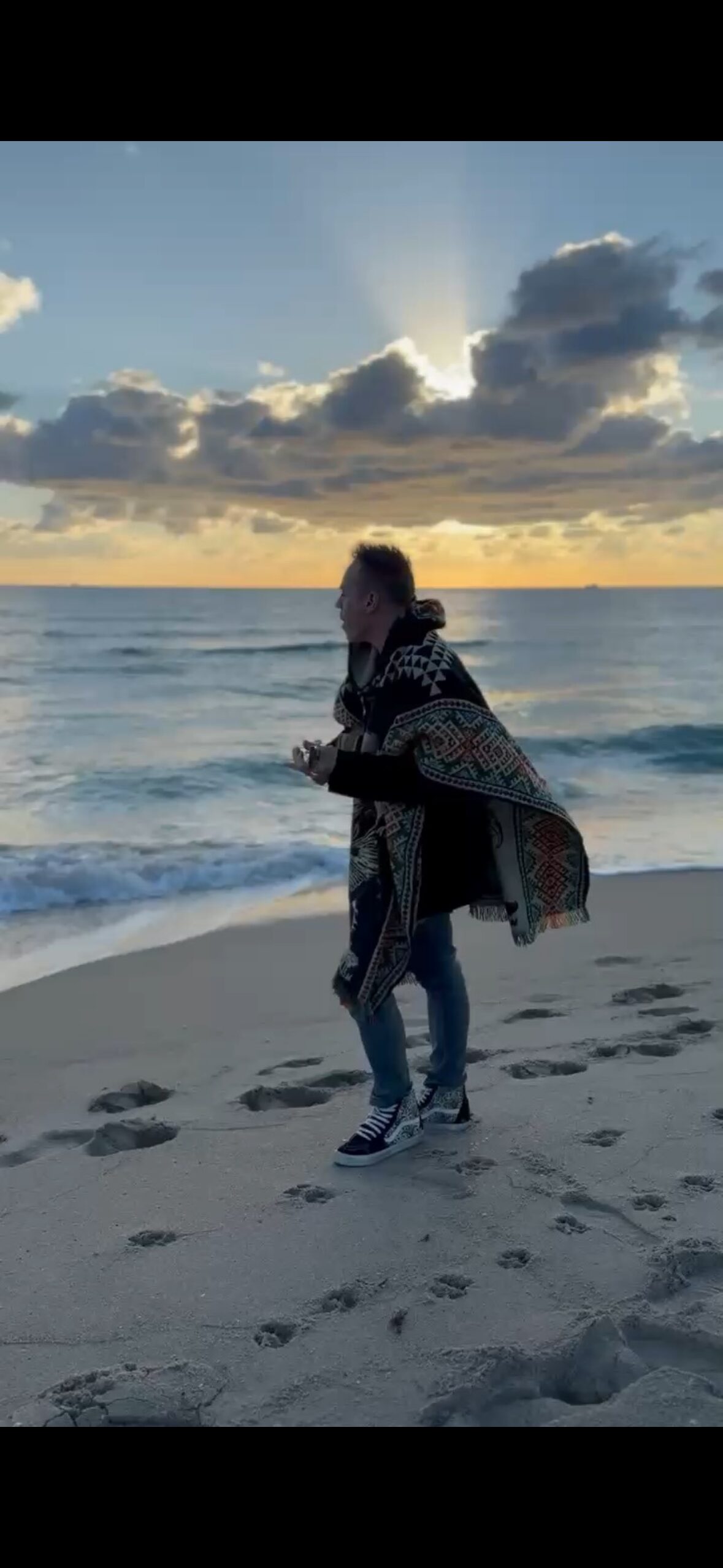Ryan on a beach at sunrise in a patterned poncho, eyes closed, singing with a ukulele toward the open water.